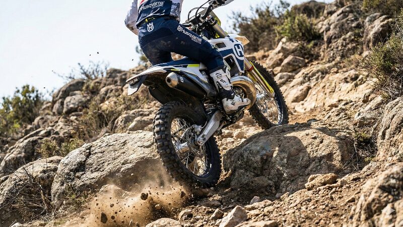 大きな岩がゴロゴロしているガレ場の急斜面を登るハスクバーナTE250iのリアビュー